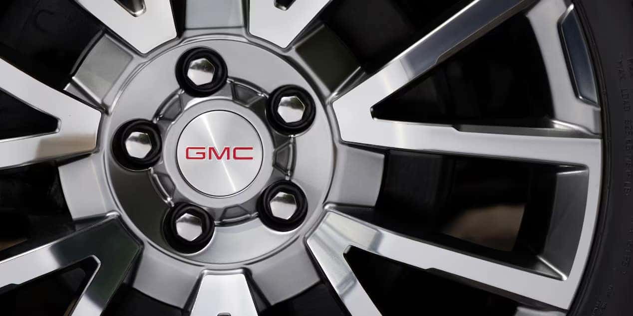 Close-up of a GMC vehicles alloy wheel with a chrome finish. The center cap displays the red GMC logo, surrounded by five lug nuts. The wheel features an intricate, modern design with spokes extending from the center.
