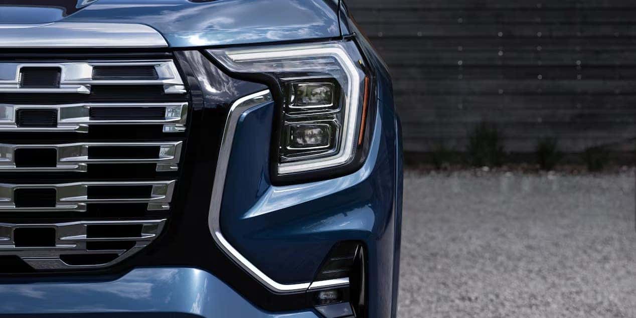 Close-up of a blue vehicles front end, focusing on the intricate grille design and angular headlight. The background is blurred, highlighting the vehicles sleek and modern aesthetic.