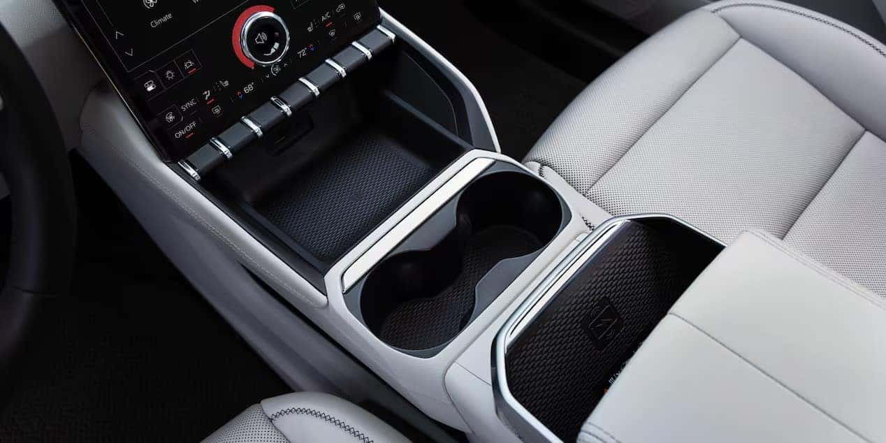 Car interior showing a center console with two cup holders, a gear selector, and a touchscreen display with various controls. The seats are upholstered in light gray leather.