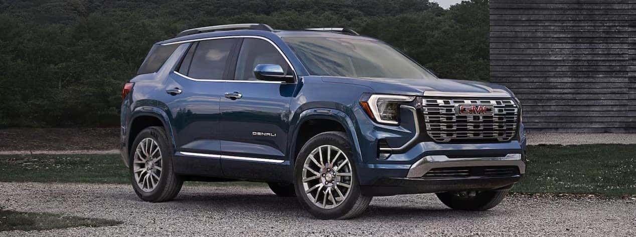 A blue SUV parked on a gravel surface in front of a wooden building. The vehicle has a chrome grille, large wheels, and tinted windows. The sky is overcast, casting a muted light on the vehicle.