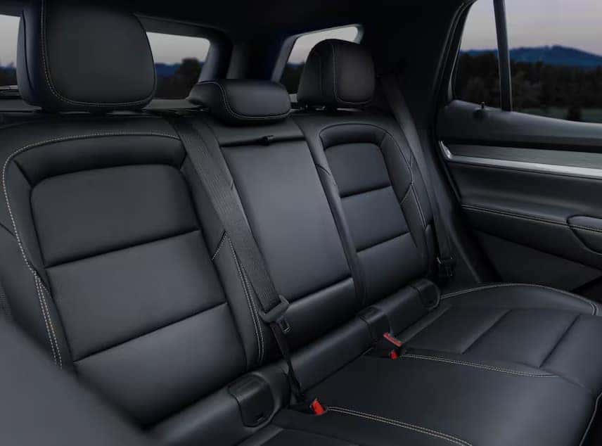 The image shows the interior of a cars backseat, featuring black leather upholstery with visible stitching. The seats have headrests and seatbelt buckles, with windows visible in the background.