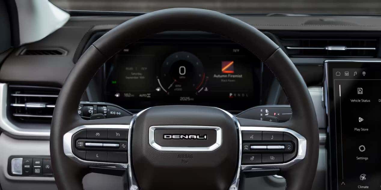 Close-up of a Denali steering wheel with visible control buttons. In the background, a digital dashboard displays speed and vehicle information, including Autumn Fermixt. The car interior features a sleek and modern black design.