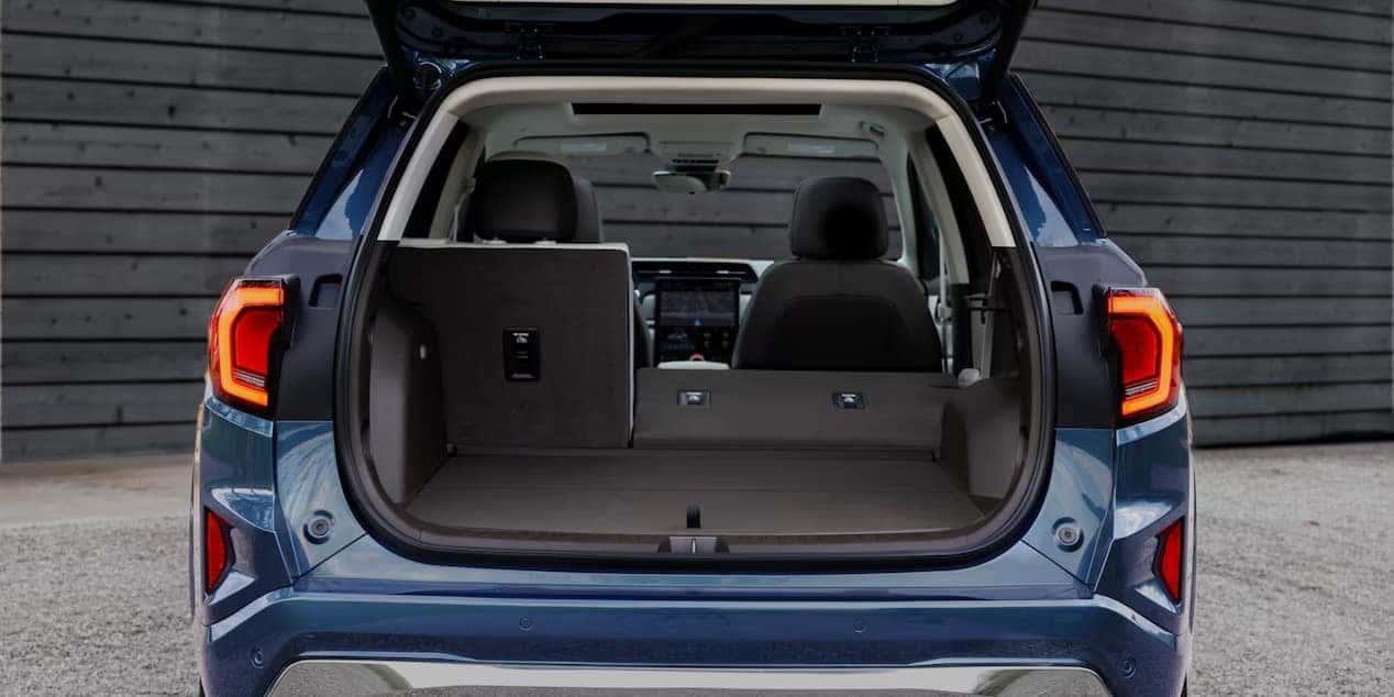 The image shows the open trunk of a blue SUV parked in front of a dark wooden wall. The back seats are partially folded, revealing a spacious cargo area. The taillights are illuminated.