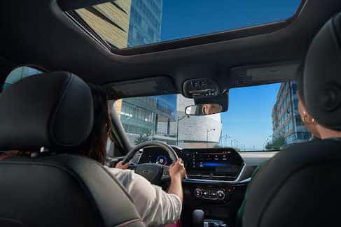 View from the backseat of a car showing two people in the front seats. The driver is holding the steering wheel. The sunroof is open, revealing a blue sky and tall buildings outside. The dashboard and rearview mirror are visible.