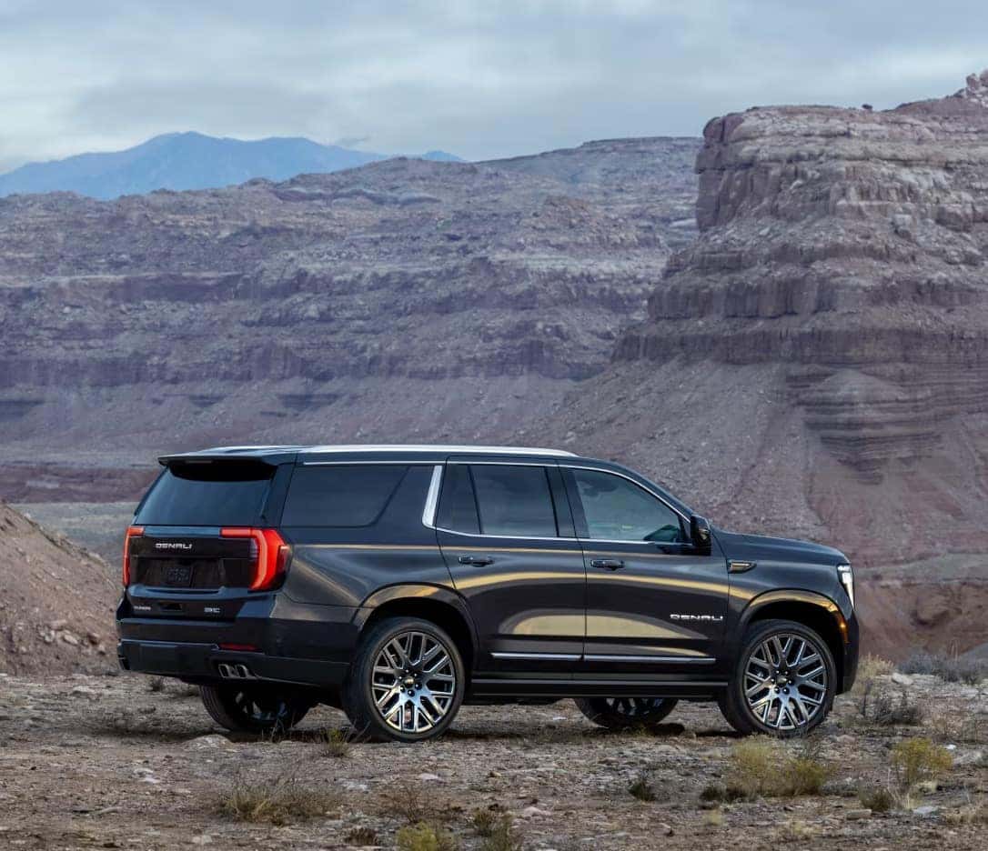 A dark SUV is parked on a rugged desert landscape with rocky formations and distant mountains under a cloudy sky. The car is viewed from the side, highlighting its sleek design and shiny wheels.