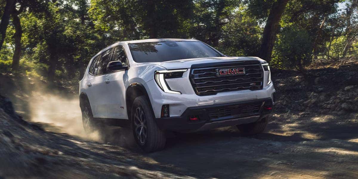 White GMC SUV driving on a dusty, rocky road surrounded by trees and greenery. The vehicle is in motion, kicking up dust, with sunlight filtering through the foliage.