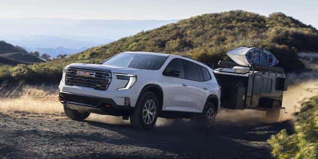 A white GMC SUV is towing a trailer on a dirt road in a hilly area. Dust is being kicked up by the vehicles movement. Hills with greenery can be seen in the background under a clear sky.