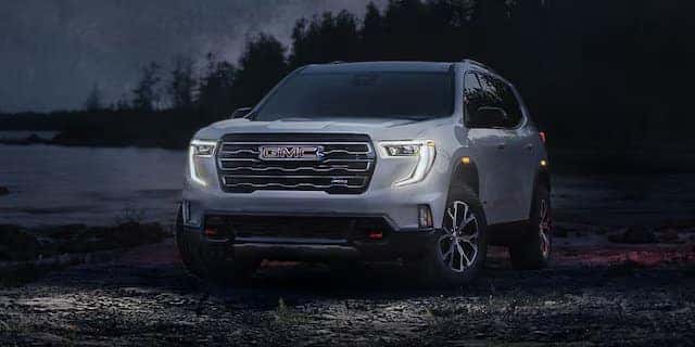 A white SUV with illuminated headlights is parked on a rocky terrain at dusk. Its surrounded by a dark, forested landscape, with the outline of trees visible in the background. The vehicles grille and branding are prominently visible.