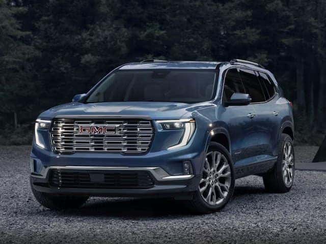 A blue GMC SUV is parked on a gravel surface, set against a backdrop of dark, blurry trees. The vehicle features a prominent front grille and sleek headlights, with a modern and polished design.