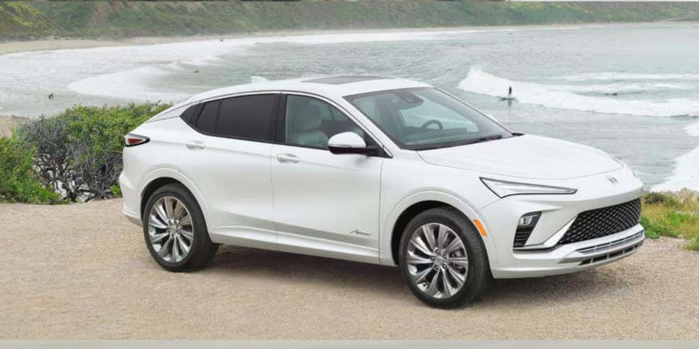 A sleek, white SUV parked on a gravel road next to a scenic coastal area. The ocean and waves are visible in the background, with surfers in the water. The car features a modern design with prominent headlights and a smooth body.