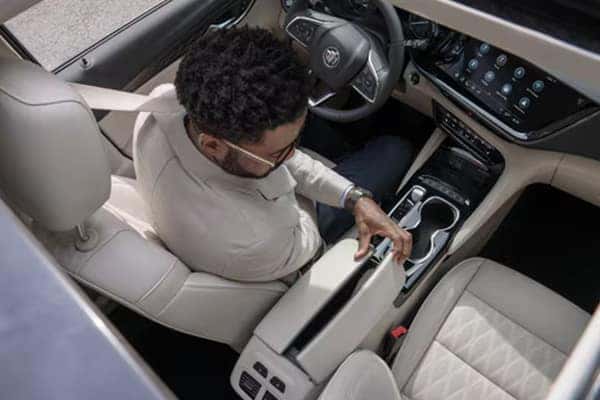 A person seated in a car adjusts the center console storage. The interior features beige seats and a touchscreen display on the dashboard. The view is from above, through the open sunroof.
