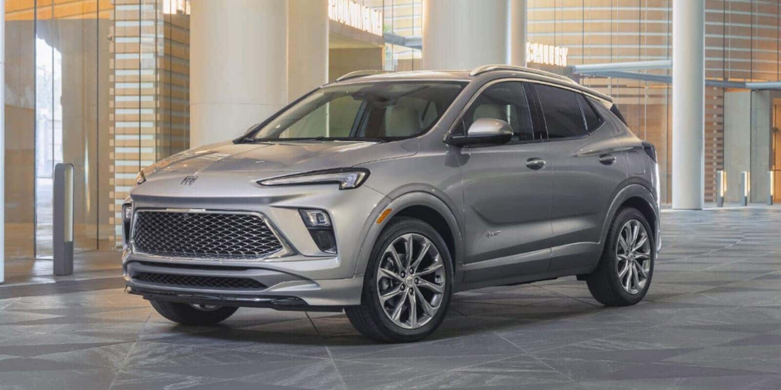 A silver SUV with sleek design and chrome accents is parked indoors on a tiled floor. It has a large grille, sharp headlights, and sporty wheels, set against a modern architectural background with columns and glass.