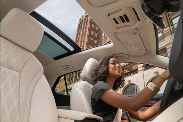 A woman is smiling while driving a car with a sunroof open. The car has white leather seats and a view of a tall building is visible through the window. She is wearing a black top and a gold bracelet.