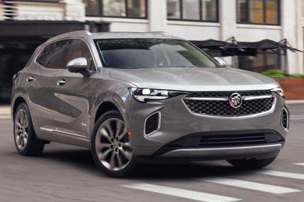 A silver SUV is driving on a city street, showing a sleek design with distinctive headlights and a prominent grille. The background features blurred buildings, suggesting motion and an urban setting.