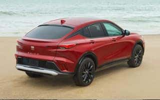 A red SUV is parked on a sandy beach near the ocean. The vehicle has a sleek, modern design with black wheels and tinted windows. The sky is overcast.
