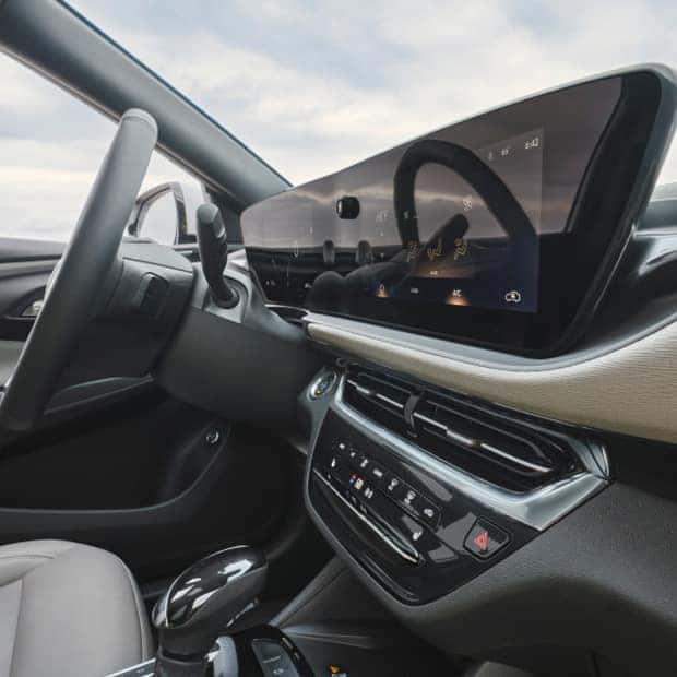 Car interior showing a sleek dashboard with a large digital display reflecting the steering wheel. The dashboard includes climate controls and other buttons. The interior features neutral tones with a gearshift in the foreground.