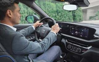 A person in a blue suit drives a car with a modern dashboard. Large display screens show various controls. Green foliage is visible through the drivers side window.