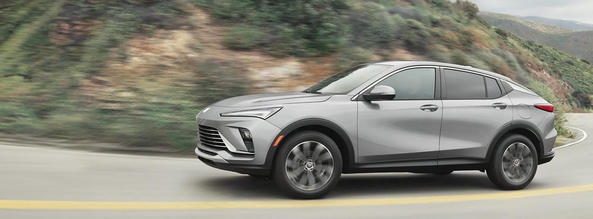 A silver SUV drives on a winding road with lush green hills in the background. The vehicle is in motion, showing its side profile as it navigates the curve. The day is clear, highlighting the sleek design of the car.