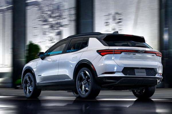 A sleek, modern white SUV is parked on a wet, reflective surface in an urban setting. The car features stylish alloy wheels and tinted windows, with the brand logo visible on the rear. Buildings with glass facades are blurred in the background.