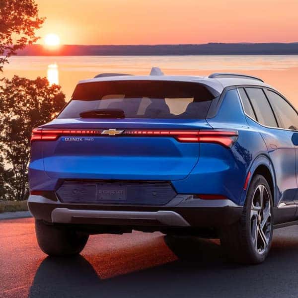 A blue SUV parked on a road, facing a large body of water during sunset. The sky is vibrant with orange and pink hues. The vehicles rear features prominent taillights, branded with a Chevrolet emblem, surrounded by lush trees.