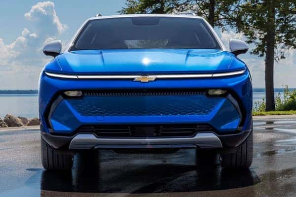 Front view of a blue Chevrolet SUV parked on a wet road near a body of water. Trees and a clear blue sky with clouds are in the background.