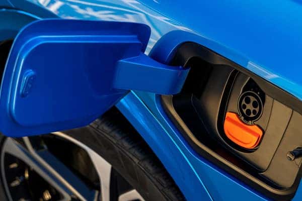 Close-up of an electric cars charging port. The vehicle is a vibrant blue, and the ports cover is open, revealing the circular connector with an orange accent. Part of a wheel is visible in the bottom left corner.