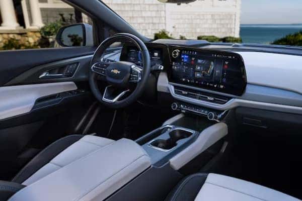 Interior of a modern car featuring a sleek dashboard with a large digital display, steering wheel with controls, and comfortable leather seats. The exterior view shows a coastline and part of a house.