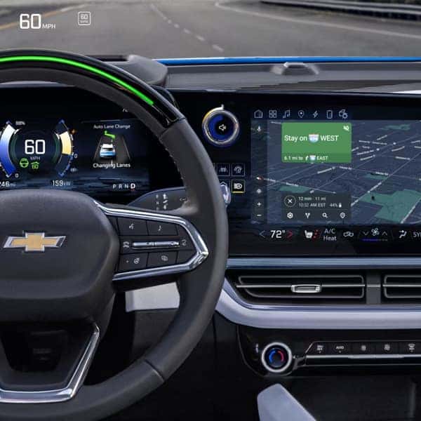 Close-up of a car dashboard with a digital speedometer showing 60 MPH. The steering wheel features a Chevrolet logo. A large touchscreen displays GPS navigation, while other controls show settings for temperature and driving mode.