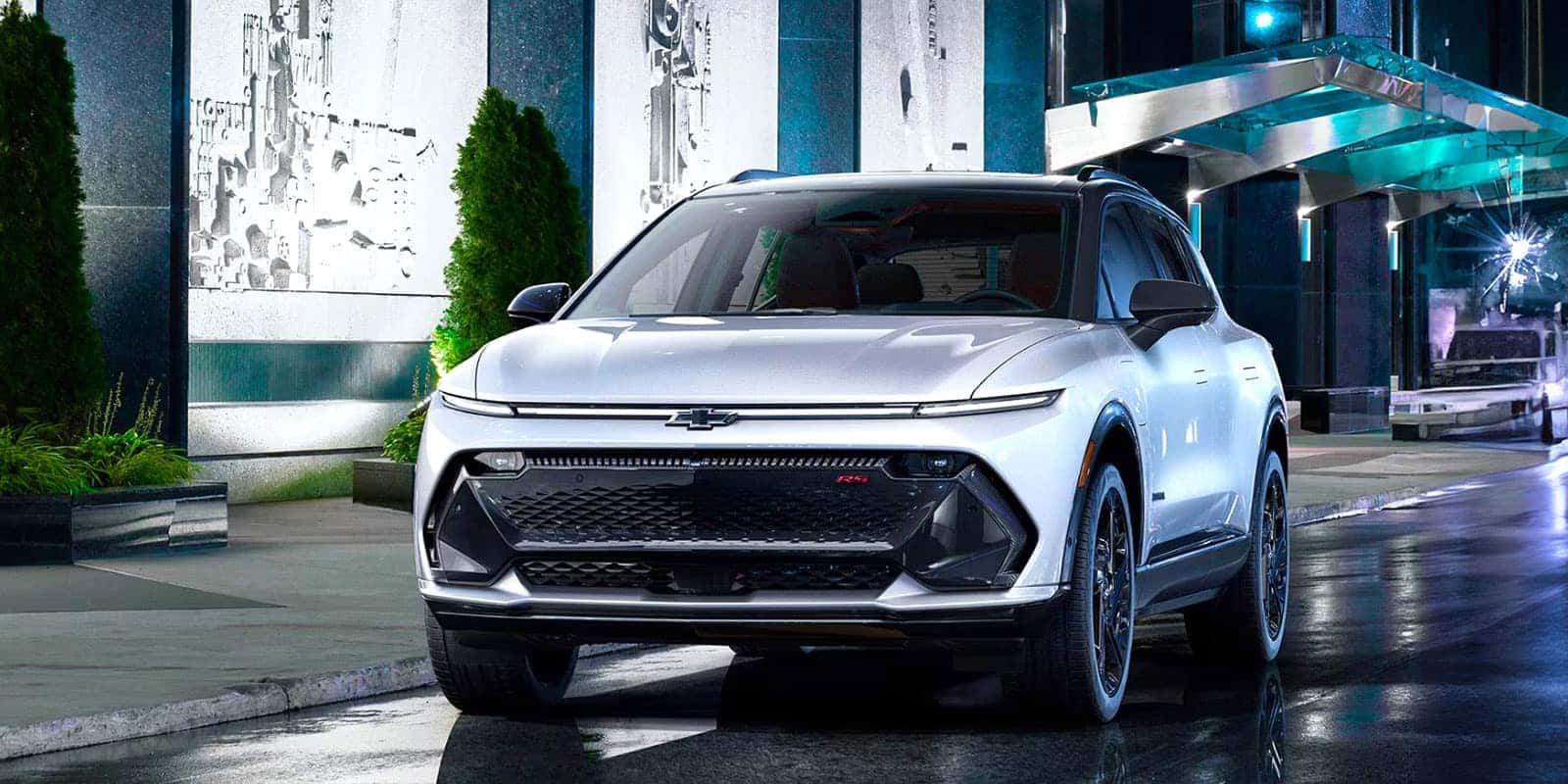 A white SUV is parked on a wet street at night in an urban setting with modern buildings. The SUV has sleek lines, distinctive headlights, and a prominent front grille. Reflections on the pavement enhance the futuristic atmosphere.
