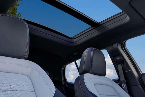 Car interior featuring two-tone seats and a large sunroof open to a blue sky with clouds. The dashboard and steering wheel are partially visible.