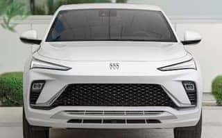 A white SUV is parked with a clear view of its front. It features sleek, narrow headlights, a prominent black grille with a geometric pattern, and a badge on the hood. The background includes greenery and a light-colored wall.
