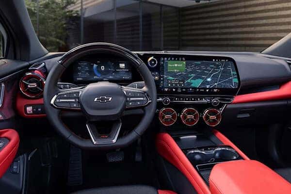 Interior view of a car showcasing a Chevrolet steering wheel, a digital dashboard, and a wide touchscreen displaying navigation. The interior features a sleek design with red and black accents, including sporty red seating and round air vents.