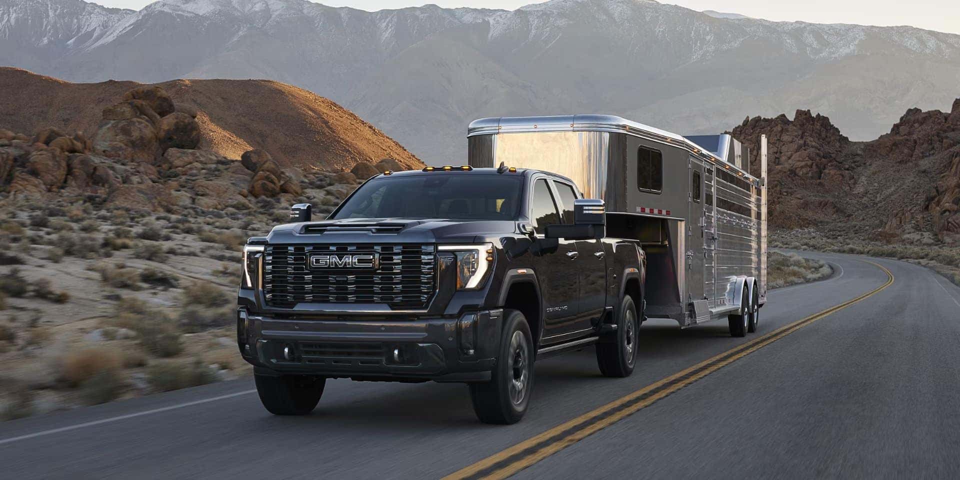 A black GMC truck is towing a silver trailer on a winding road through a desert landscape. Mountains and rocky terrain are visible in the background under a clear sky.