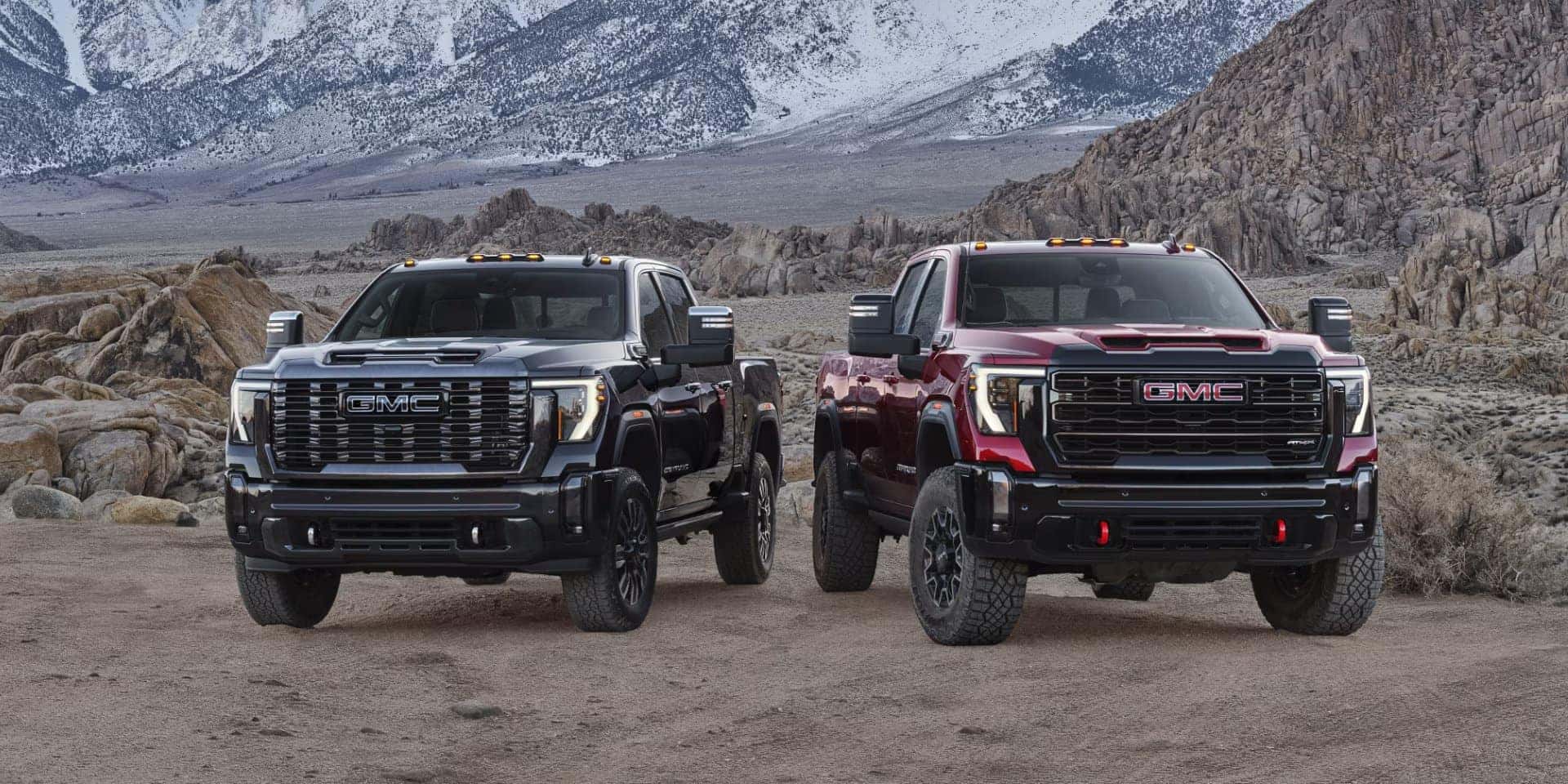 Two GMC pickup trucks, one black and one red, are parked on a rugged landscape with rocky terrain and snow-capped mountains in the background. Both trucks have a robust, off-road design with raised suspensions and prominent front grills.