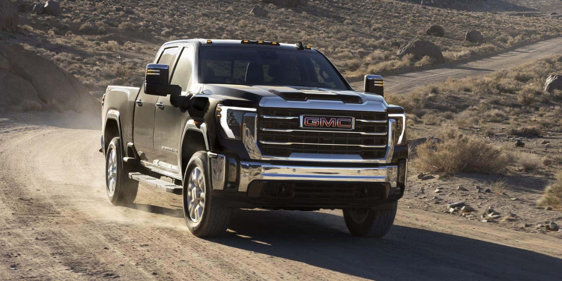 A black GMC pickup truck is driving on a dusty, desert trail. The landscape is arid with sparse vegetation and rocky hills in the background. The truck kicks up dust as it moves along the rugged path.