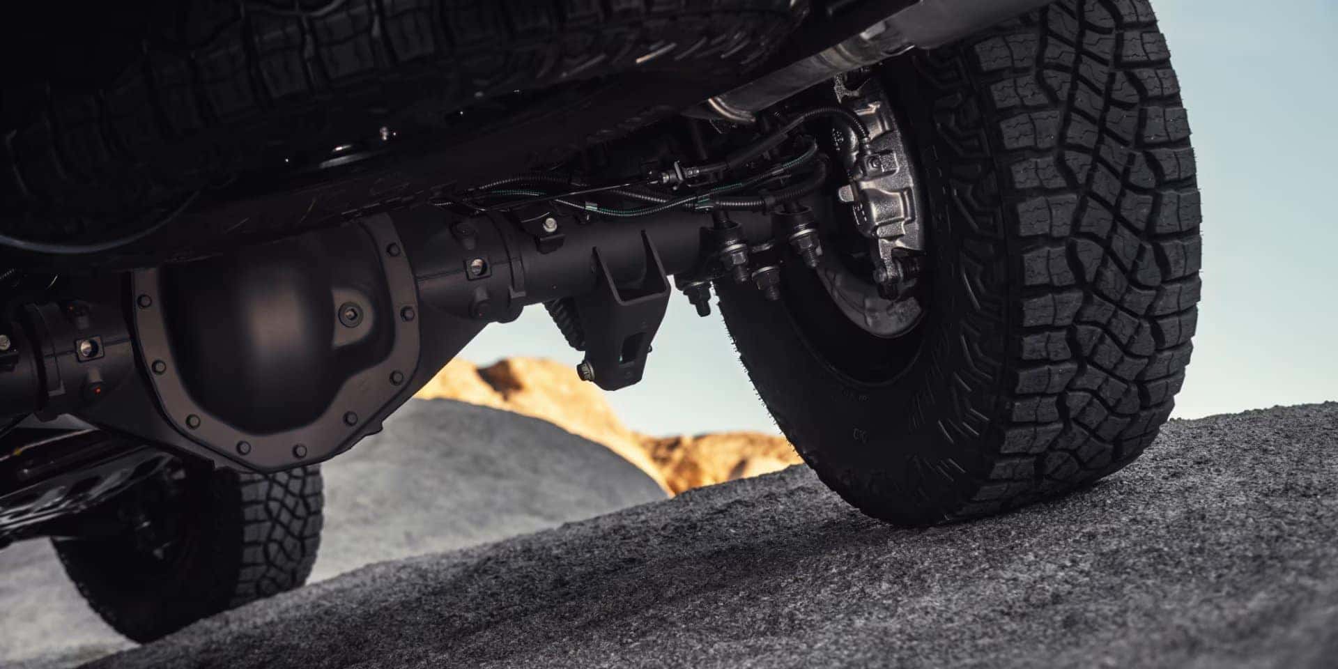 Close-up view of a vehicles underside with rugged tires positioned on rocky terrain. A mountain is visible in the background under a clear sky, emphasizing off-road capability.