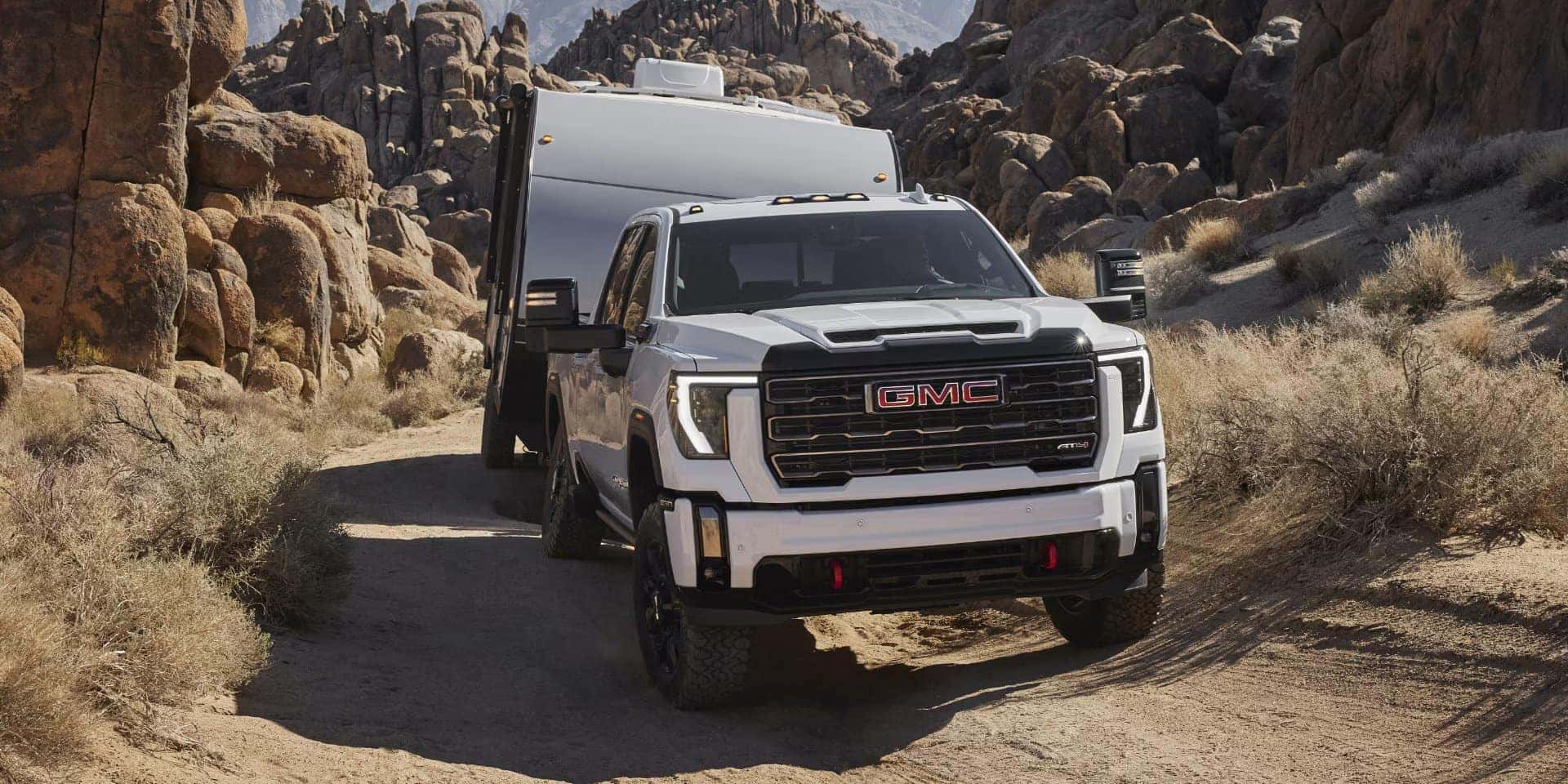 A GMC truck towing a large white trailer navigates a narrow, rocky desert path. The background features rugged, sandy-colored rock formations under a clear sky. The trucks lights are on, highlighting its powerful build and design.