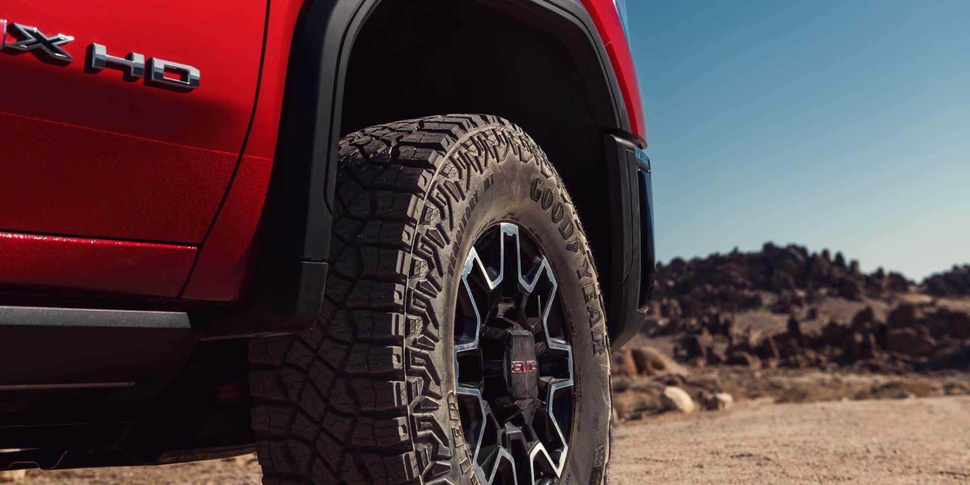 Close-up of a red off-road truck with large, rugged Goodyear tires, parked on a gravel terrain under a clear blue sky. Rocky hills are visible in the background.