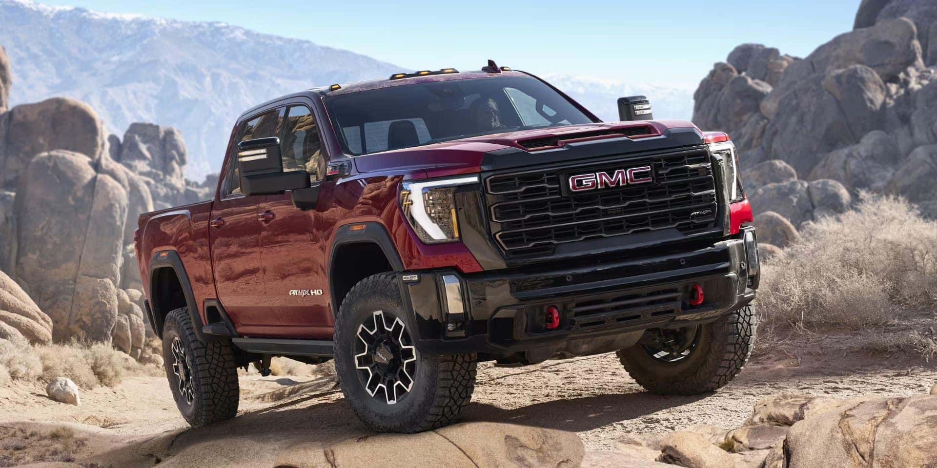A red GMC pickup truck on a rocky terrain with mountains in the background under a clear sky. The truck features a robust front grille and off-road tires.