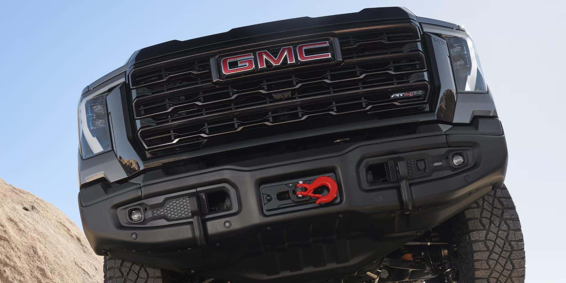 Low-angle view of a black GMC trucks front, showcasing its bold grille, red GMC logo, and front bumper with a red tow hook. The truck is positioned on a rocky terrain, reflecting an off-road setting against a clear sky.