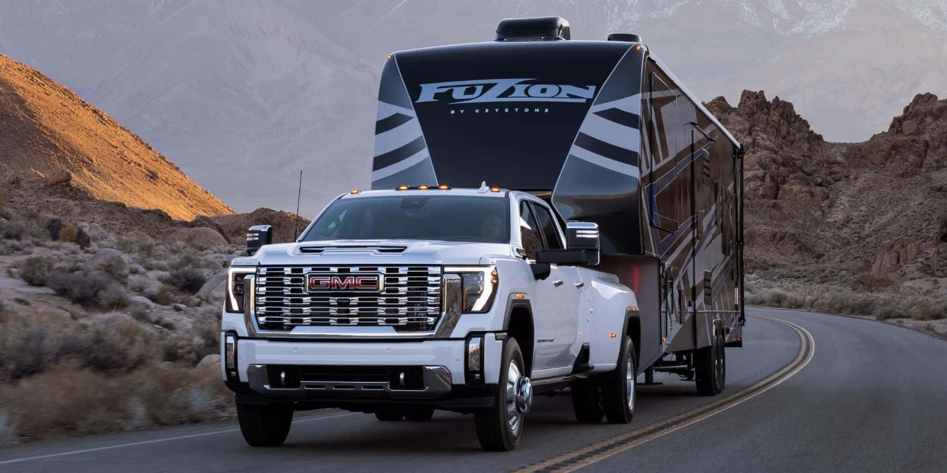 A white GMC truck towing a large black and gray RV labeled Fuzion on a winding road through a desert landscape with rocky hills in the background. The sky is overcast, casting a muted light over the scene.