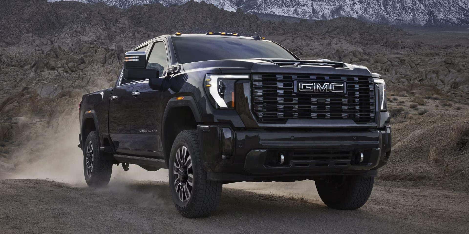A black GMC pickup truck drives on a dusty, rugged terrain with rocky hills in the background. Snow-covered mountains are visible in the distance under a cloudy sky. The trucks headlights are on.