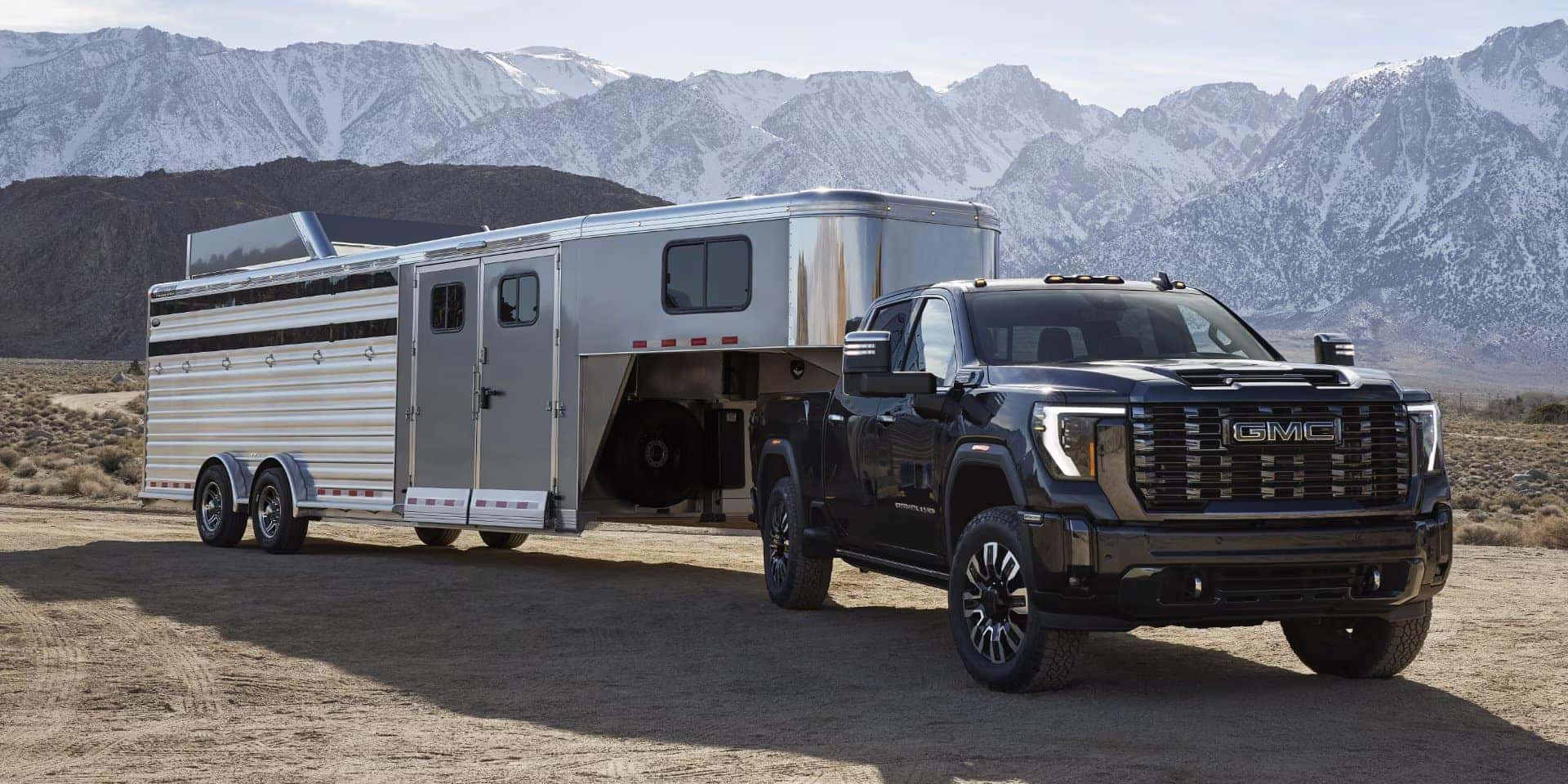 A black GMC pickup truck towing a large, silver trailer is parked on a dirt road. Snow-capped mountains and a clear sky form the background, creating a scenic, rugged landscape.