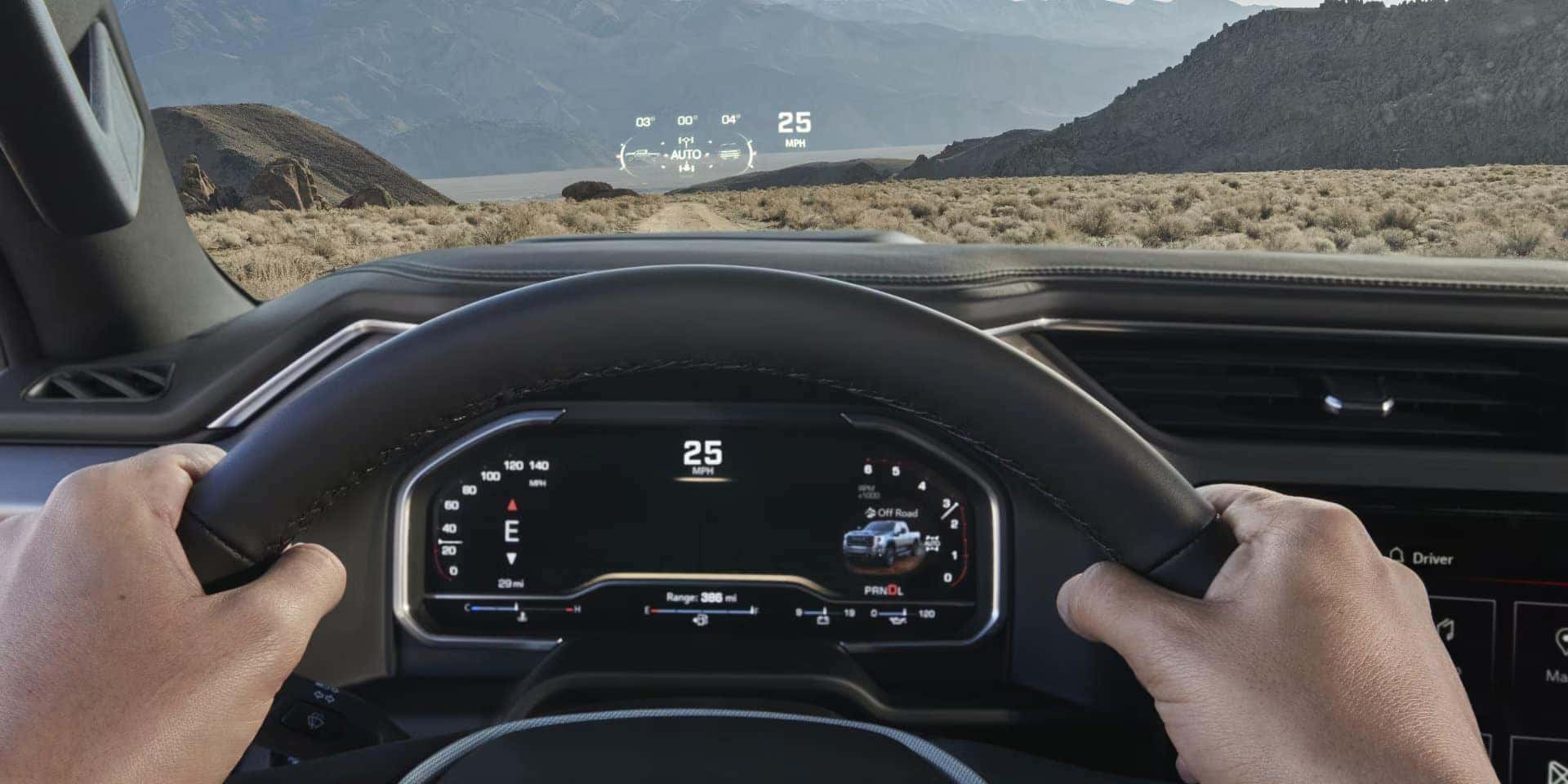 The image shows a person driving a vehicle on a rugged terrain, viewing through the windshield. The dashboard displays speed and vehicle stats. A heads-up display projects information onto the windshield, with mountains visible in the distance.