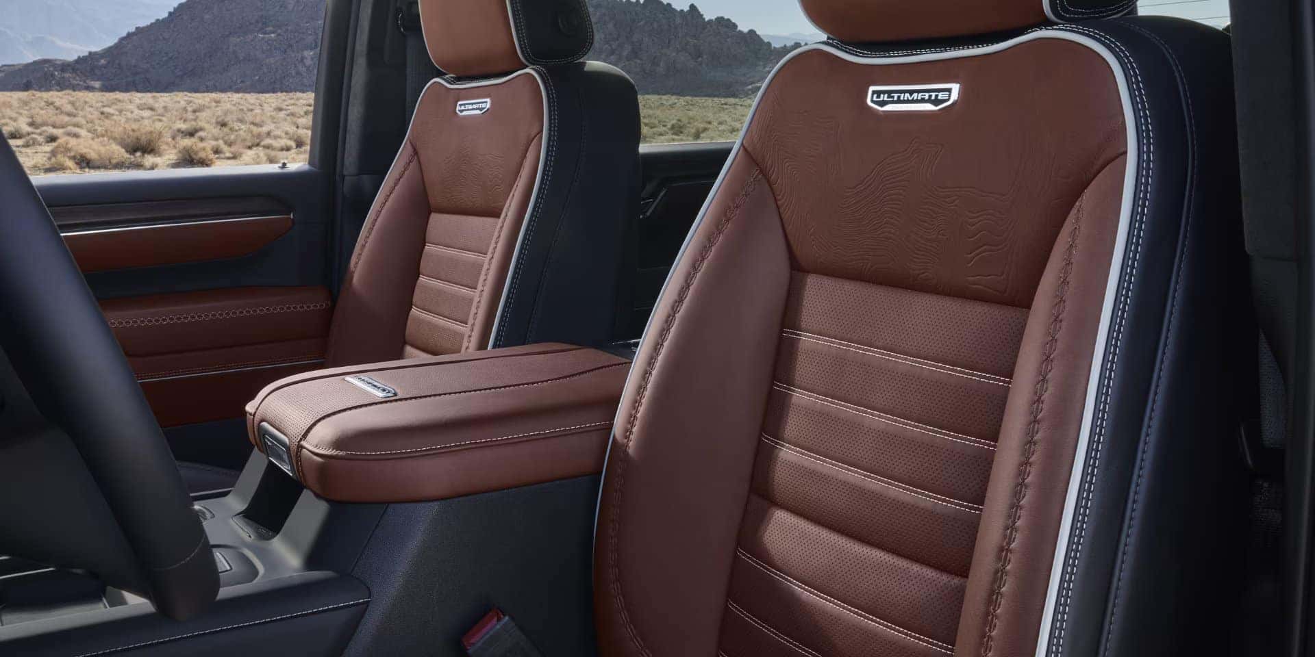 The image shows the interior of a vehicle, featuring two front seats with brown leather upholstery and detailed stitching. The headrests have the word ULTIMATE embroidered on them. The background shows a desert landscape through the window.