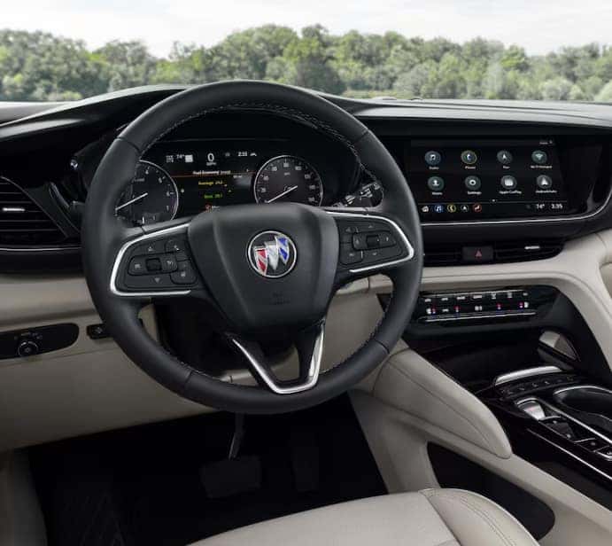 The image shows the interior of a car, focusing on the steering wheel with a Buick logo. The dashboard displays digital gauges and a touchscreen with various apps. The interior features leather and modern controls. Trees are visible through the windshield.