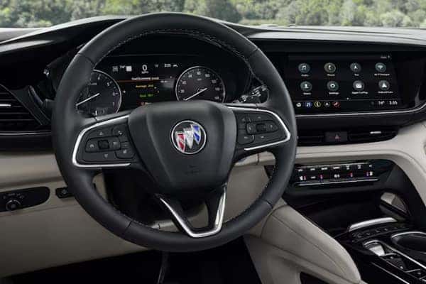 Close-up of a car dashboard featuring a black steering wheel with the Buick logo. Behind it, there are analog speedometer and tachometer gauges. To the right, a digital display shows various controls. Forested landscape visible through the windshield.