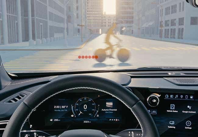 View from inside a car showing a cyclist crossing the street at an intersection in a city. The dashboard displays controls and information. Sunlight and shadows create a clear, bright scene.