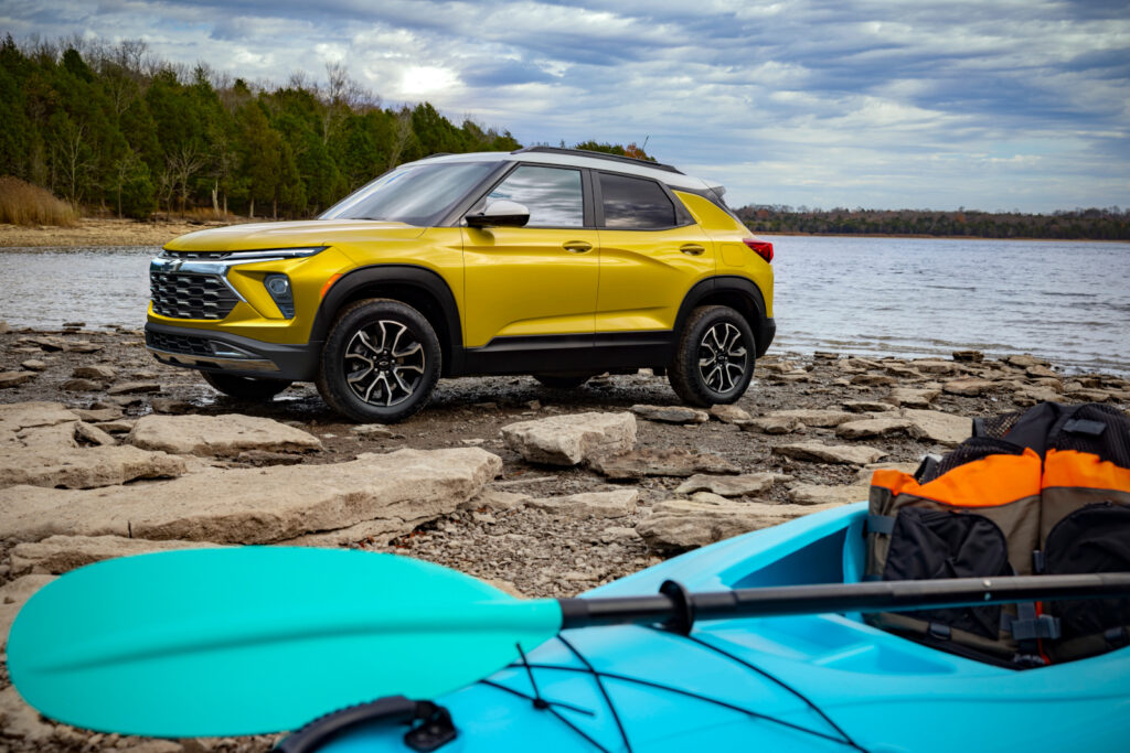 Yellow SUV parked on rocky lakeside with a blue kayak and paddles in the foreground. Vibrant outdoor scene.