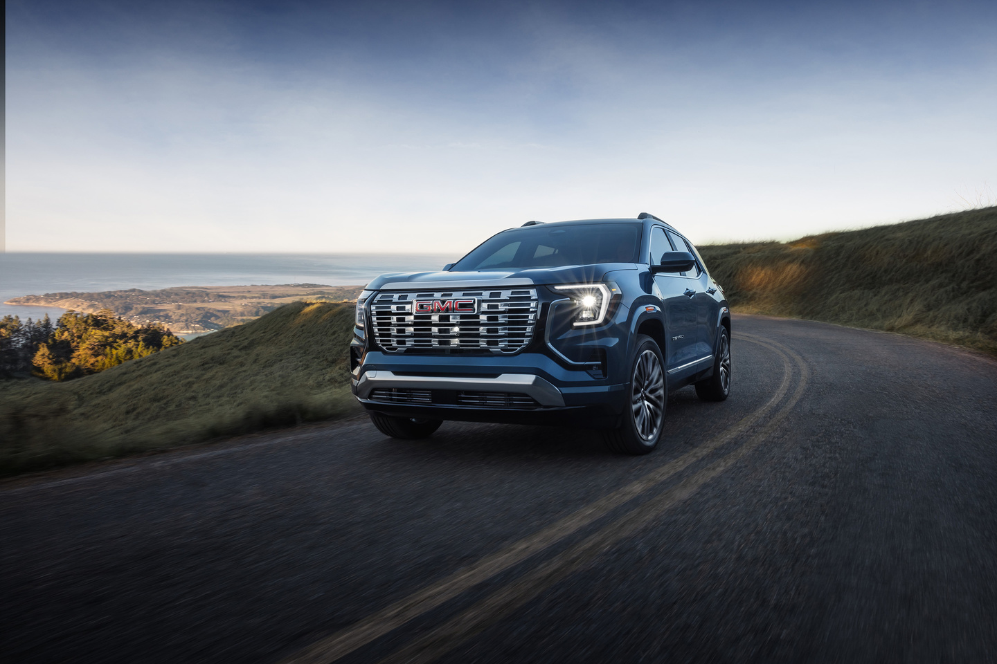 A blue GMC SUV drives on a scenic coastal road, with hills and ocean in the background under a clear sky.